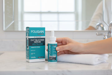 มือกำลังถือเซรั่ม DHT FOLIGAIN ข้างกล่องและผ้าขนหนูที่พับอยู่บนเคาน์เตอร์ห้องน้ำ.
