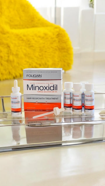 กล่องและขวดไมน็อกซิดิล FOLIGAIN บนชั้นในห้องน้ำข้างผ้าขนหนูสีเหลือง.