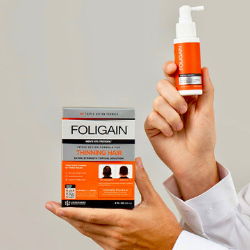 มือกำลังถือกล่องและขวดสเปรย์ผลิตภัณฑ์บำรุงชนิดทา TRIOXIDIL 10% FOLIGAIN สำหรับผู้ชาย.