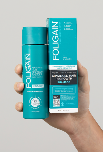 มือกำลังถือแชมพูสำหรับการงอกใหม่ของเส้นผมสูตรแอดวานซ์ FOLIGAIN ในขวดสีเขียวอมฟ้าข้างกล่อง.